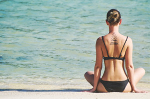 Eine Frau mit einer Tätowierung im Nacken sitzt am Strand, blickt aufs Meer und meditiert.